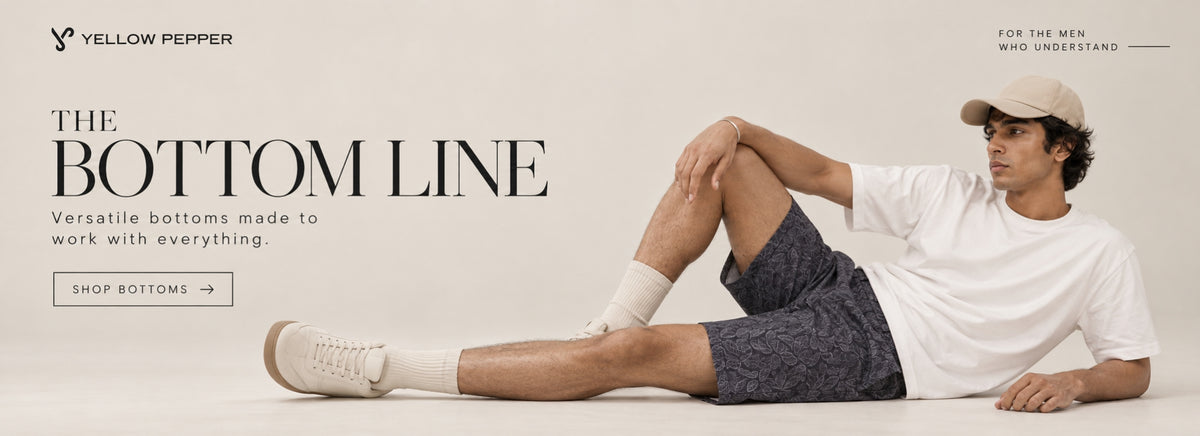A man wearing a white t-shirt, dark patterned shorts, white socks, and beige sneakers sits on the floor against a light beige studio background. The image features promotional text for 'Yellow Pepper' about 'The Bottom Line' and includes a 'Shop Bottoms' button.