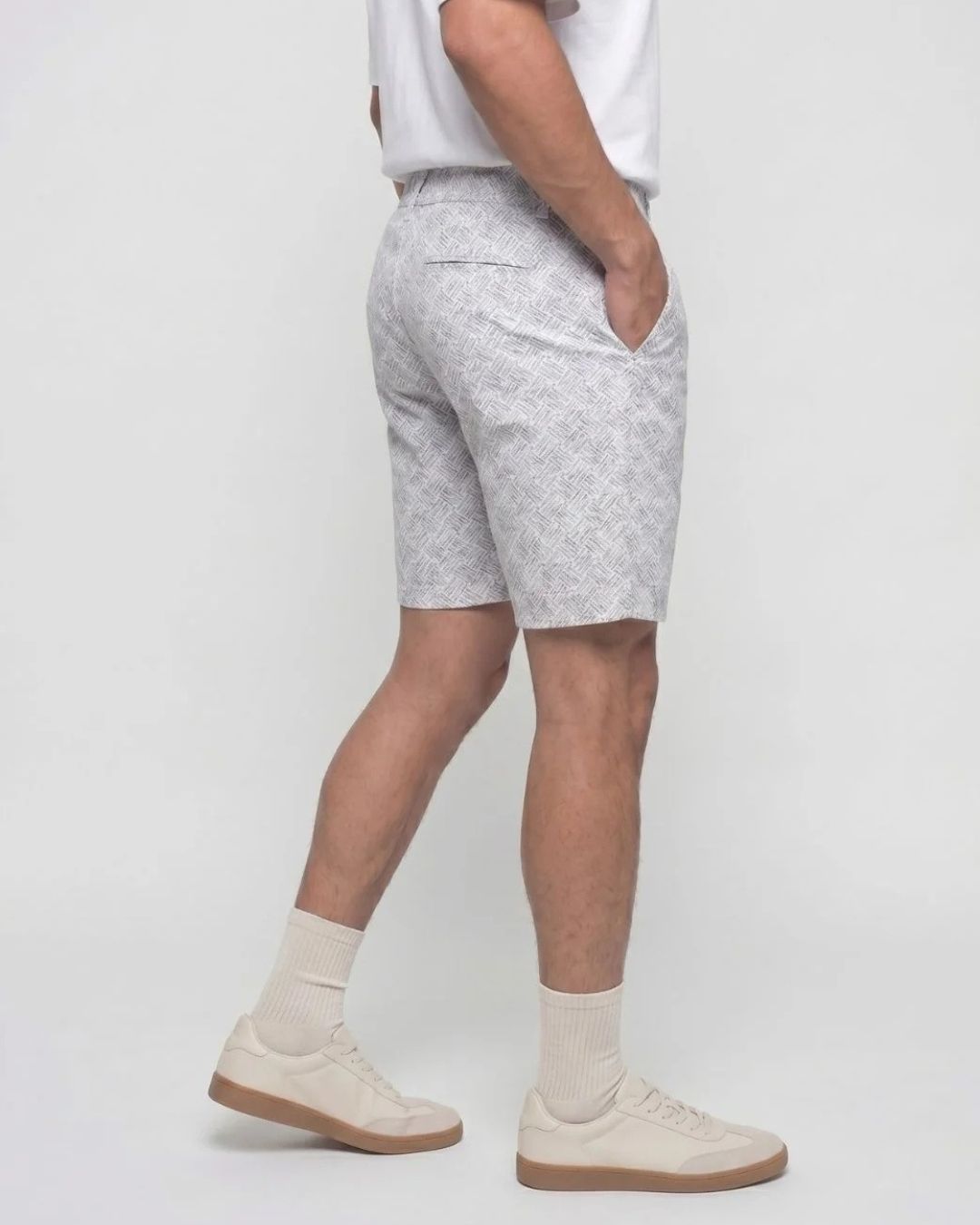 A person shown from the waist down wearing light gray patterned shorts, cream socks, and cream sneakers against a plain light gray studio background.