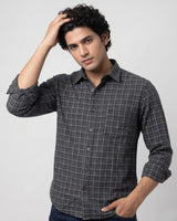 A man wearing a dark gray and black plaid button-down shirt, posing with one hand on his hair against a light gray studio background.