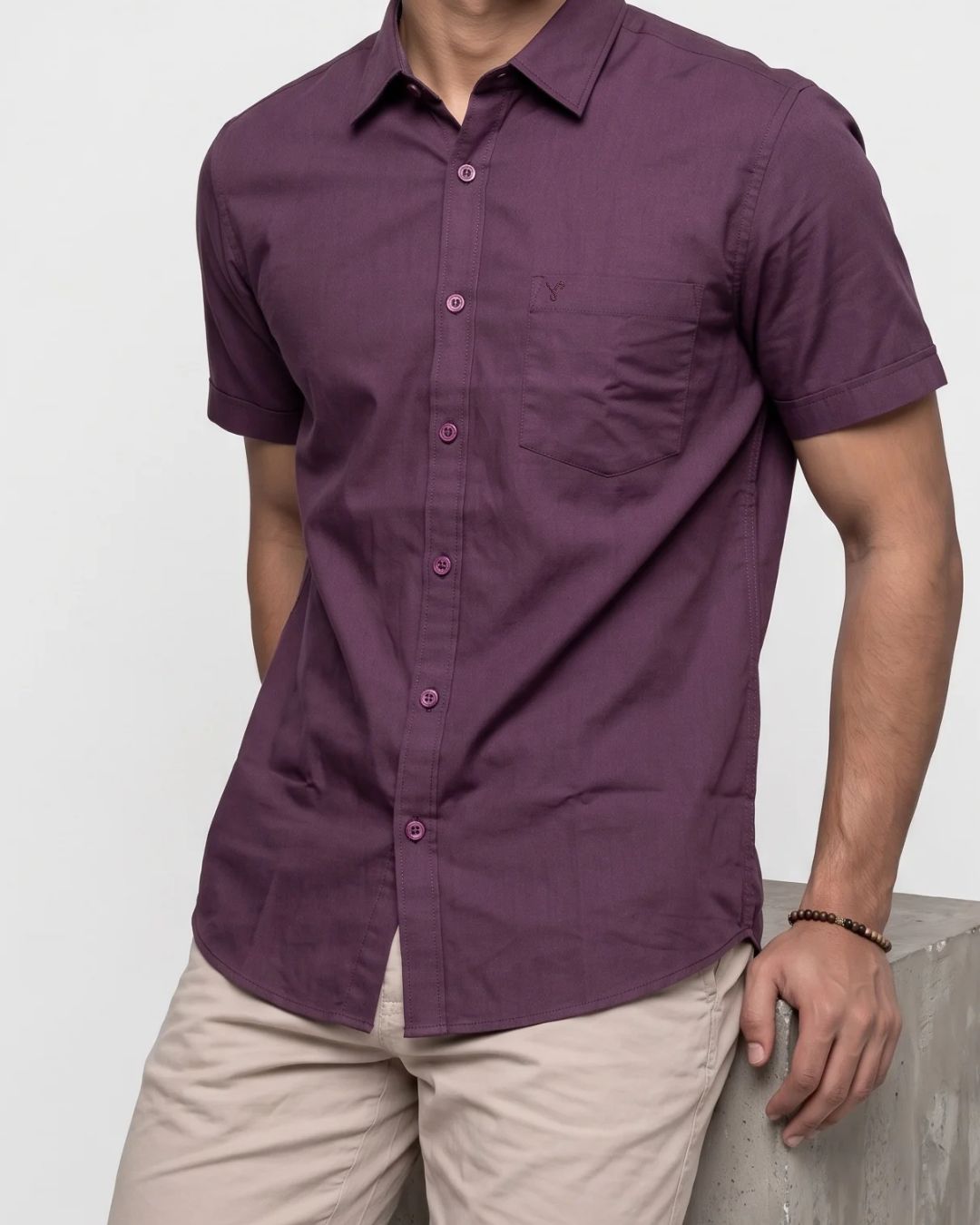 A man wearing a short-sleeve button-up shirt in a deep plum purple color, paired with beige trousers, standing against a light gray background.