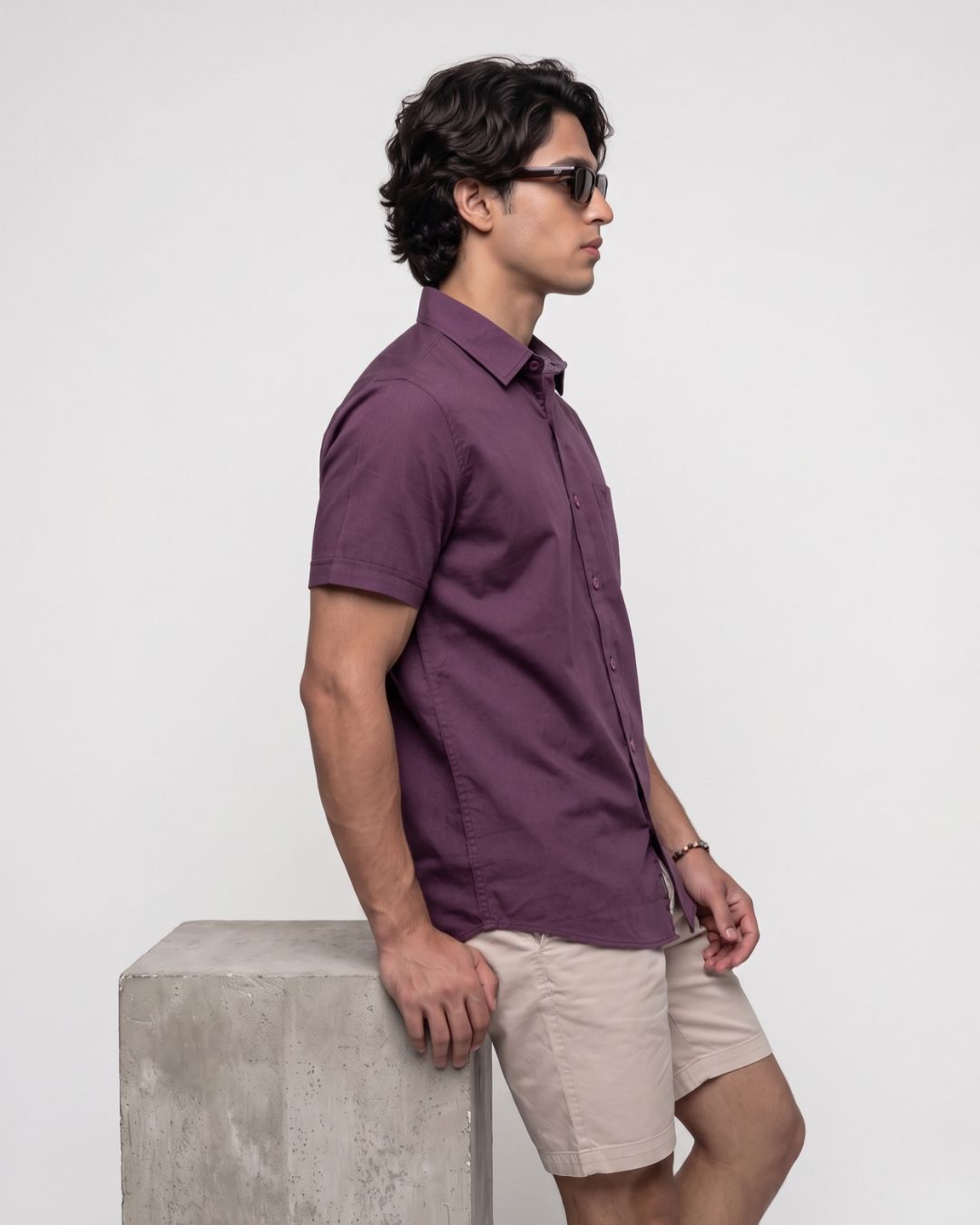 A man wearing a short-sleeve button-up shirt in a deep plum purple and beige shorts, standing in profile next to a concrete block against a light gray background.