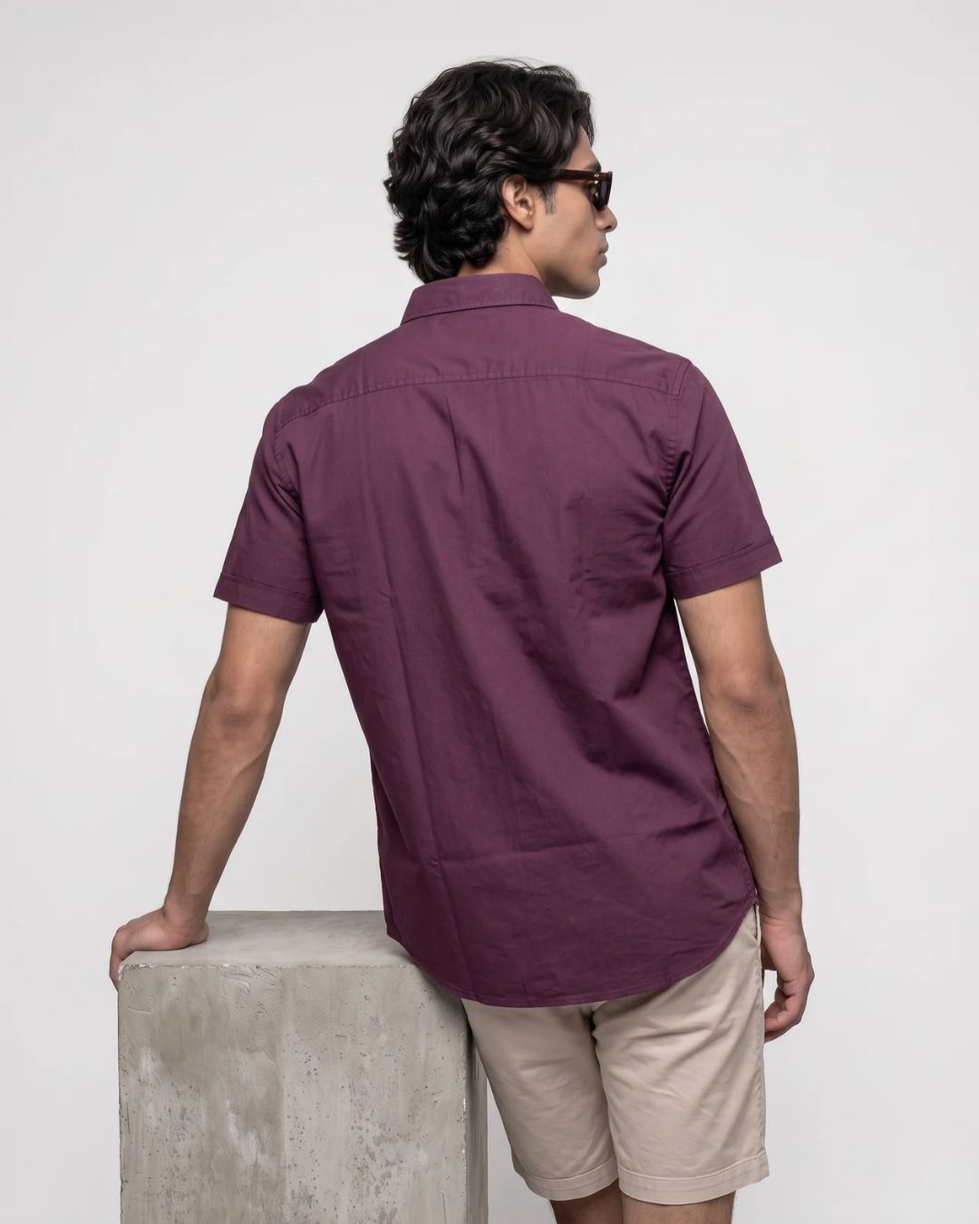 A man wearing a short-sleeve button-up shirt in a deep plum purple color, shown from the back as he leans on a concrete block against a plain white background.