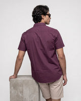 A man wearing a short-sleeve button-up shirt in a deep plum purple color, shown from the back as he leans on a concrete block against a plain white background.
