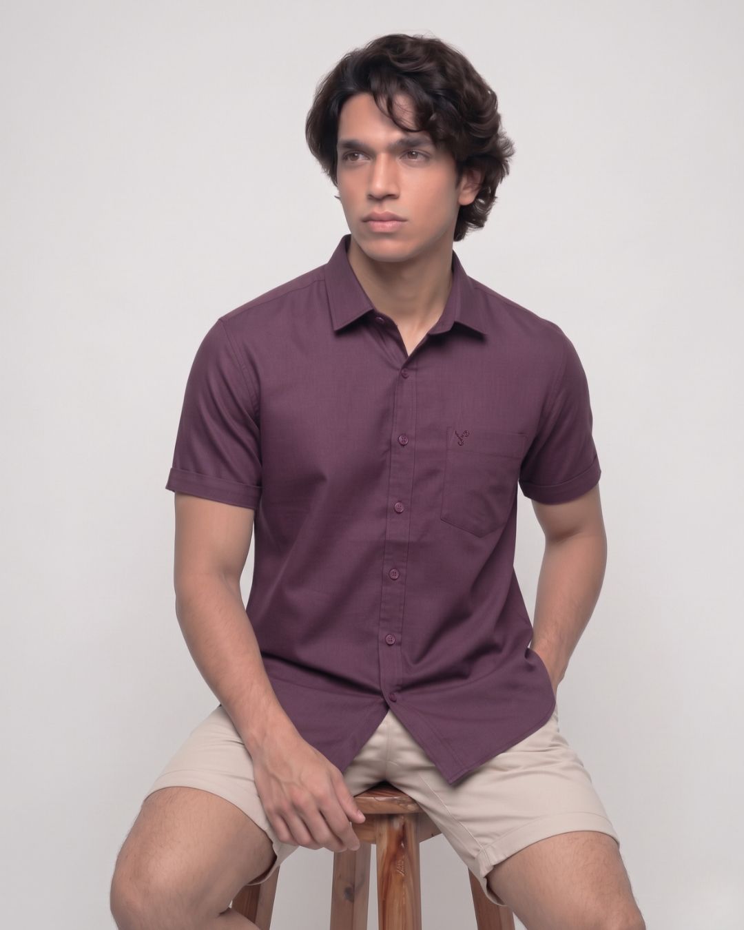 A man wearing a short-sleeve button-up shirt in a deep plum purple color, paired with beige shorts, sitting on a wooden stool against a light gray background.