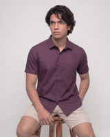 A man wearing a short-sleeve button-up shirt in a deep plum purple color, paired with beige shorts, sitting on a wooden stool against a light gray background.