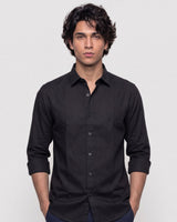 A man wearing a black long-sleeve button-down shirt with rolled-up sleeves, standing against a light gray studio background.