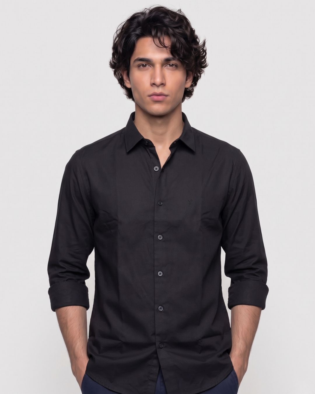 A man wearing a black long-sleeve button-down shirt with rolled-up sleeves, standing against a light gray studio background.