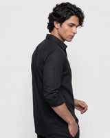 A man wearing a black long-sleeve button-down shirt, shown from a side profile against a plain light gray studio background.