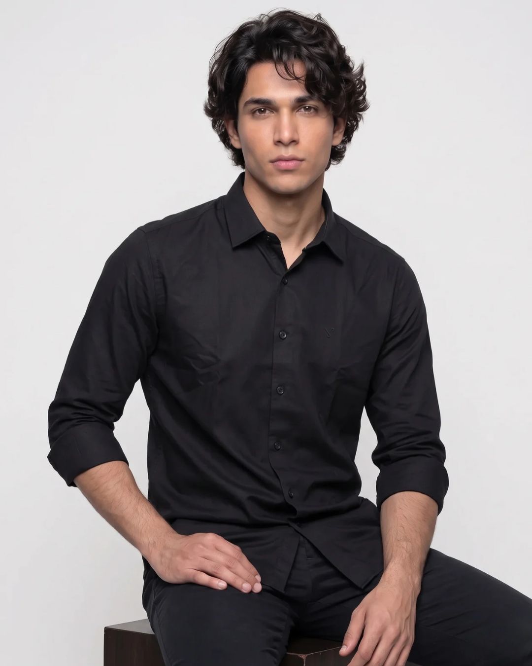 A man wearing a black long-sleeve button-up shirt and black pants, seated against a plain light gray studio background.