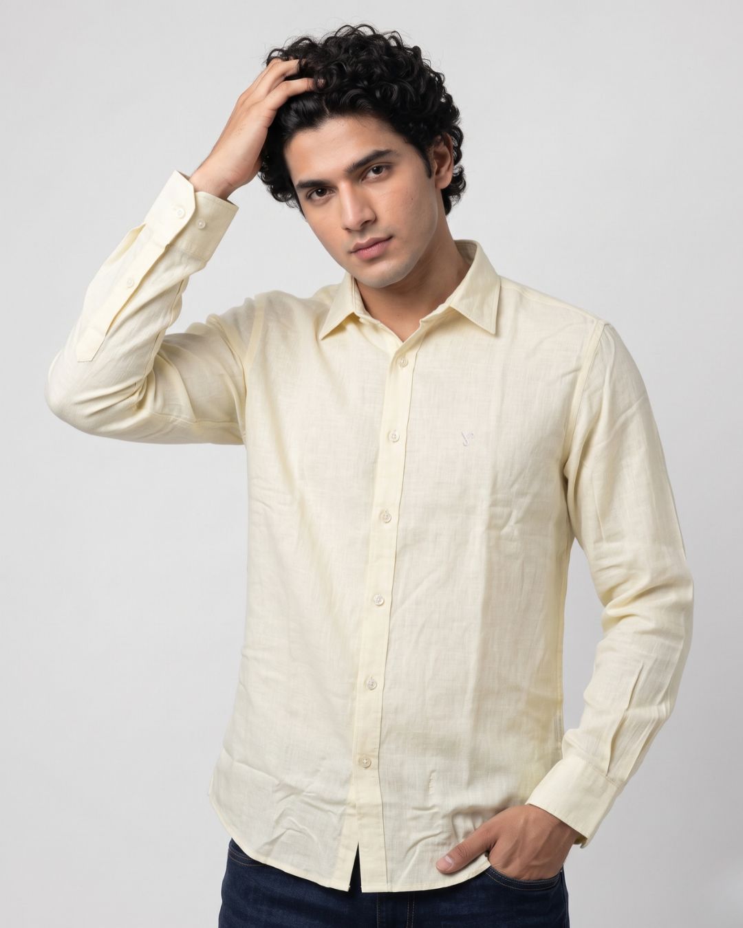 A man wearing a light cream-colored linen button-down shirt and dark blue jeans, posing against a light gray studio background.