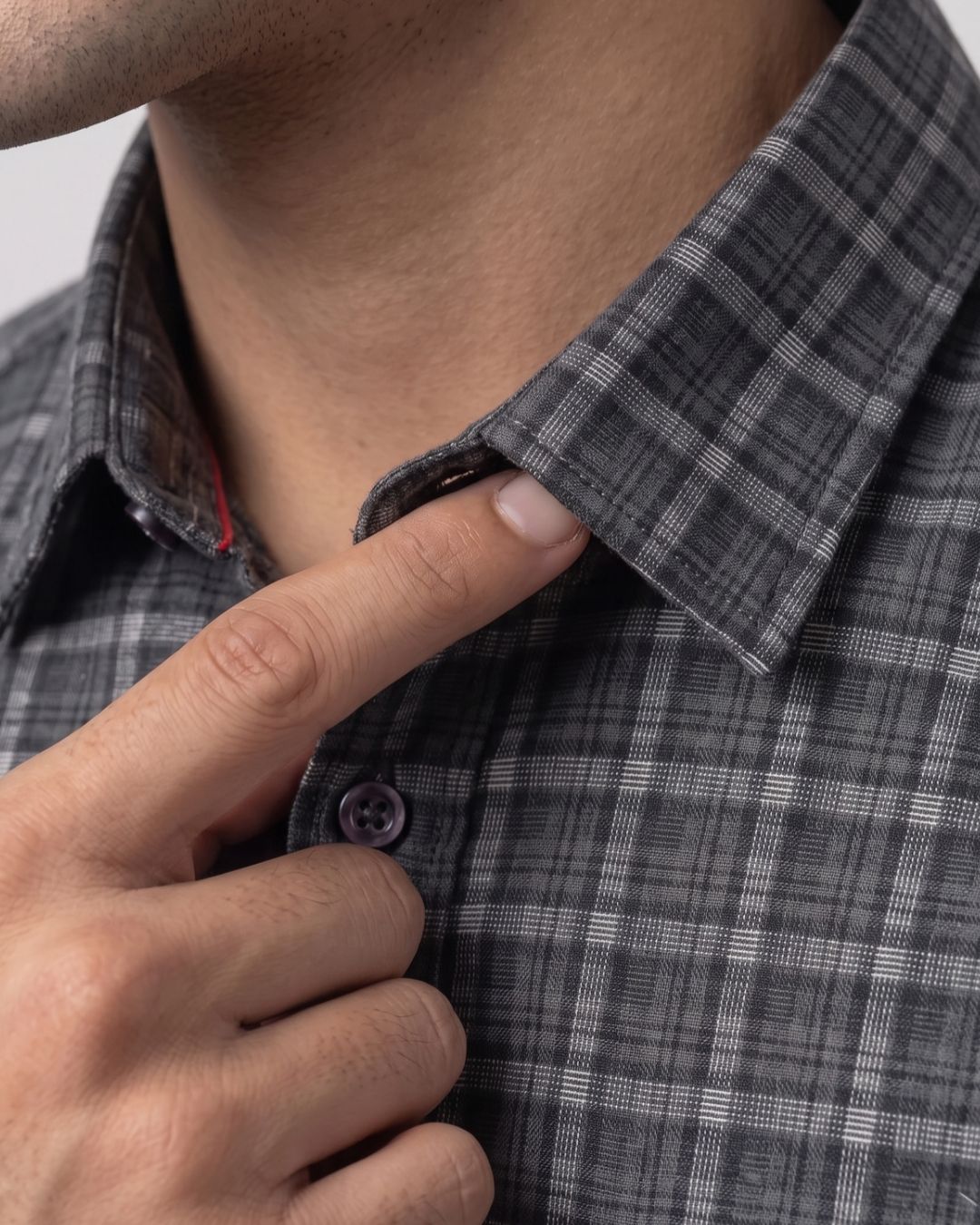 Close-up of a man wearing a dark gray and white plaid button-down shirt, adjusting the collar with his right hand.