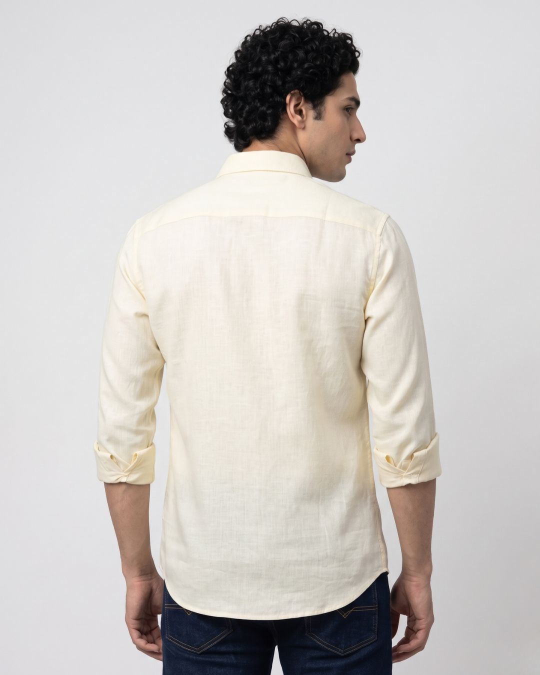 A man viewed from the back wearing a light cream-colored linen shirt with rolled-up sleeves and dark blue jeans, set against a plain light gray studio background.