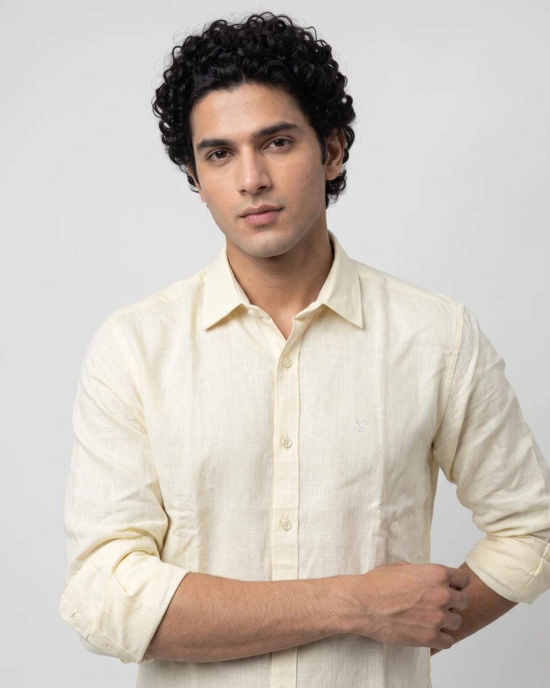 A man with curly black hair wearing a light cream-colored linen button-up shirt, photographed against a neutral light gray background.