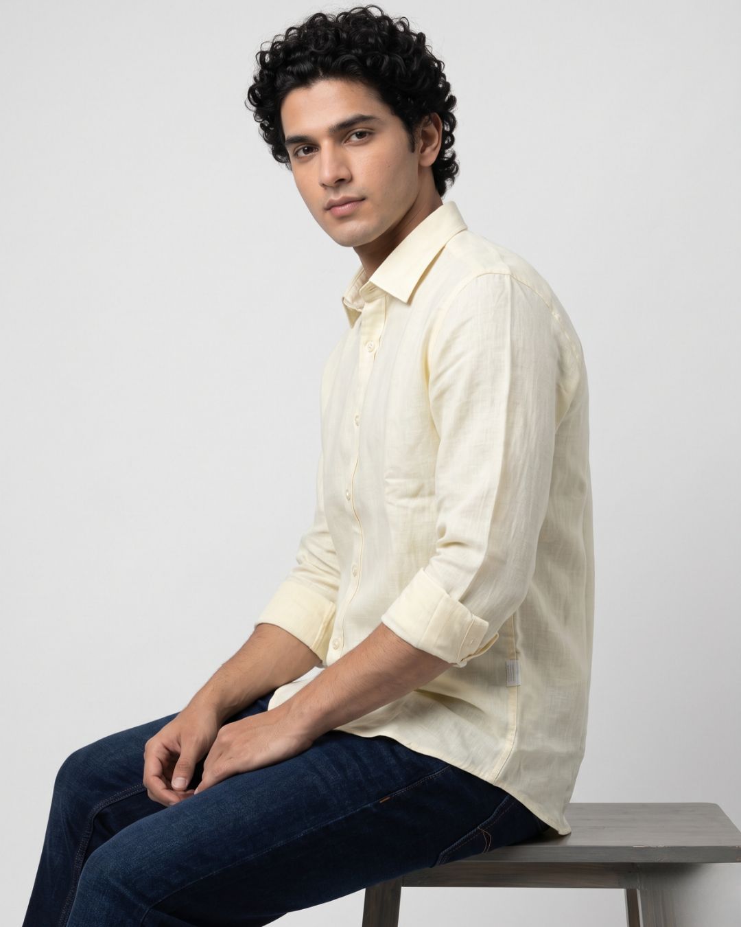A man wearing a light cream-colored long-sleeve linen shirt and dark blue jeans, sitting on a gray bench against a light gray background.