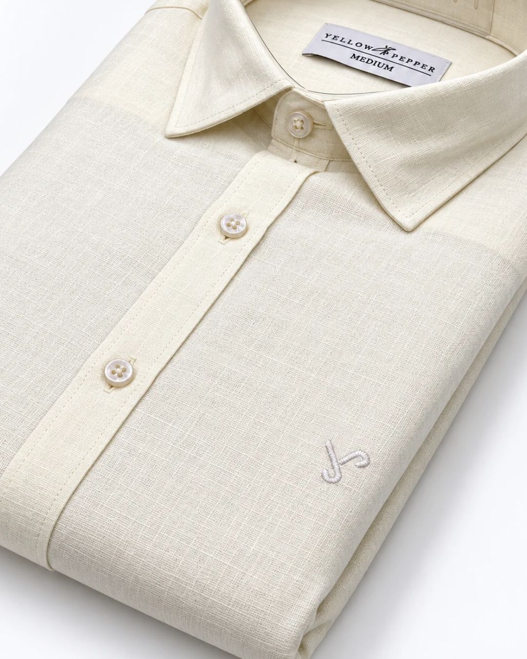 A folded cream-colored linen shirt by Yellow Pepper, featuring a textured weave, a classic collar, and a small embroidered logo on the chest.