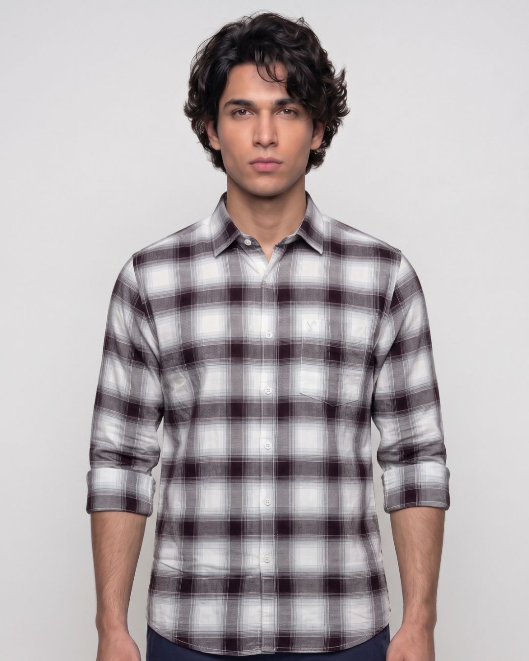 A man wearing a long-sleeve button-up plaid shirt in white, gray, and dark brown, standing against a light gray background.