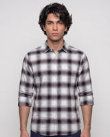 A man wearing a long-sleeve button-up plaid shirt in white, gray, and dark brown, standing against a light gray background.