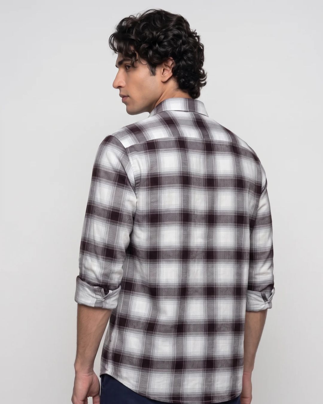 A man wearing a white long-sleeve plaid button-down shirt with dark brown and black checkered lines, shown from a three-quarter back view against a light gray background.