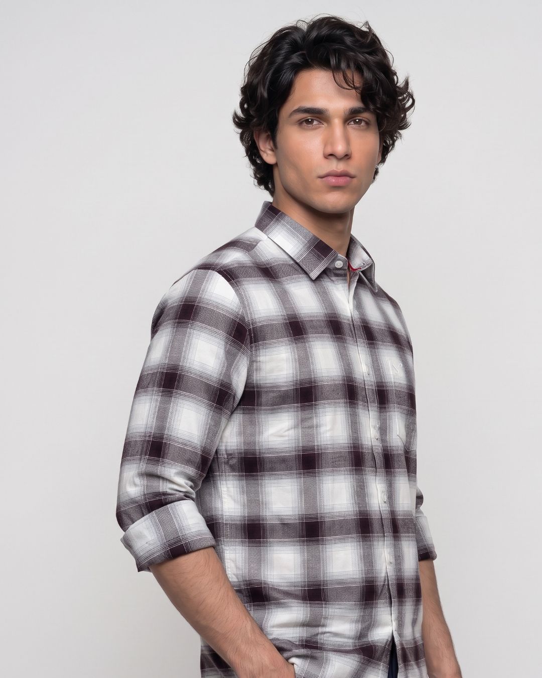 A man wearing a long-sleeve plaid button-up shirt in white, gray, and dark brown, photographed against a light gray studio background.