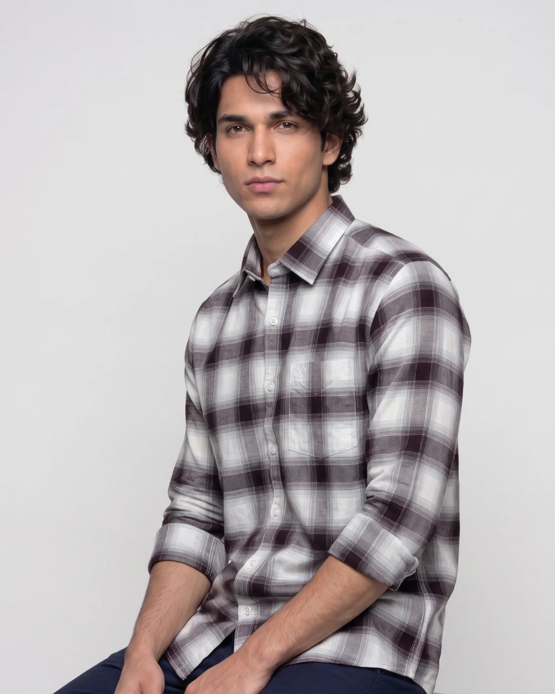 A man wearing a long-sleeve plaid button-up shirt in white, gray, and dark brown, seated against a plain light gray studio background.