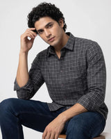 A man wearing a dark gray and black plaid button-down shirt with blue jeans, sitting against a light gray studio background.