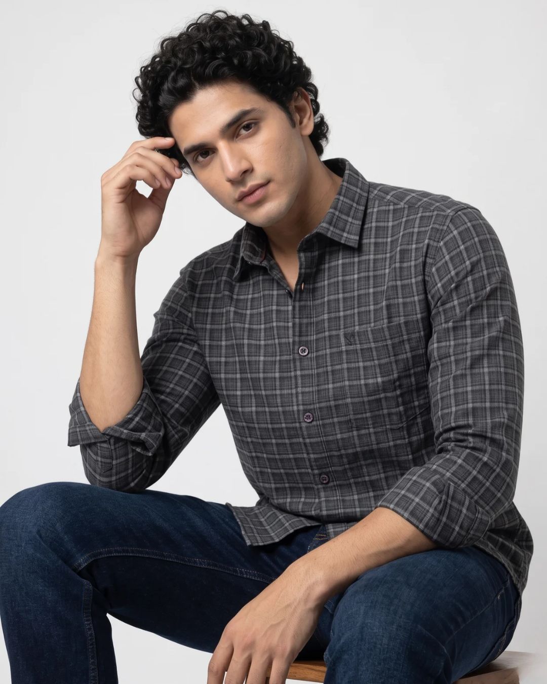 A man wearing a dark gray and black plaid button-down shirt with blue jeans, sitting against a light gray studio background.