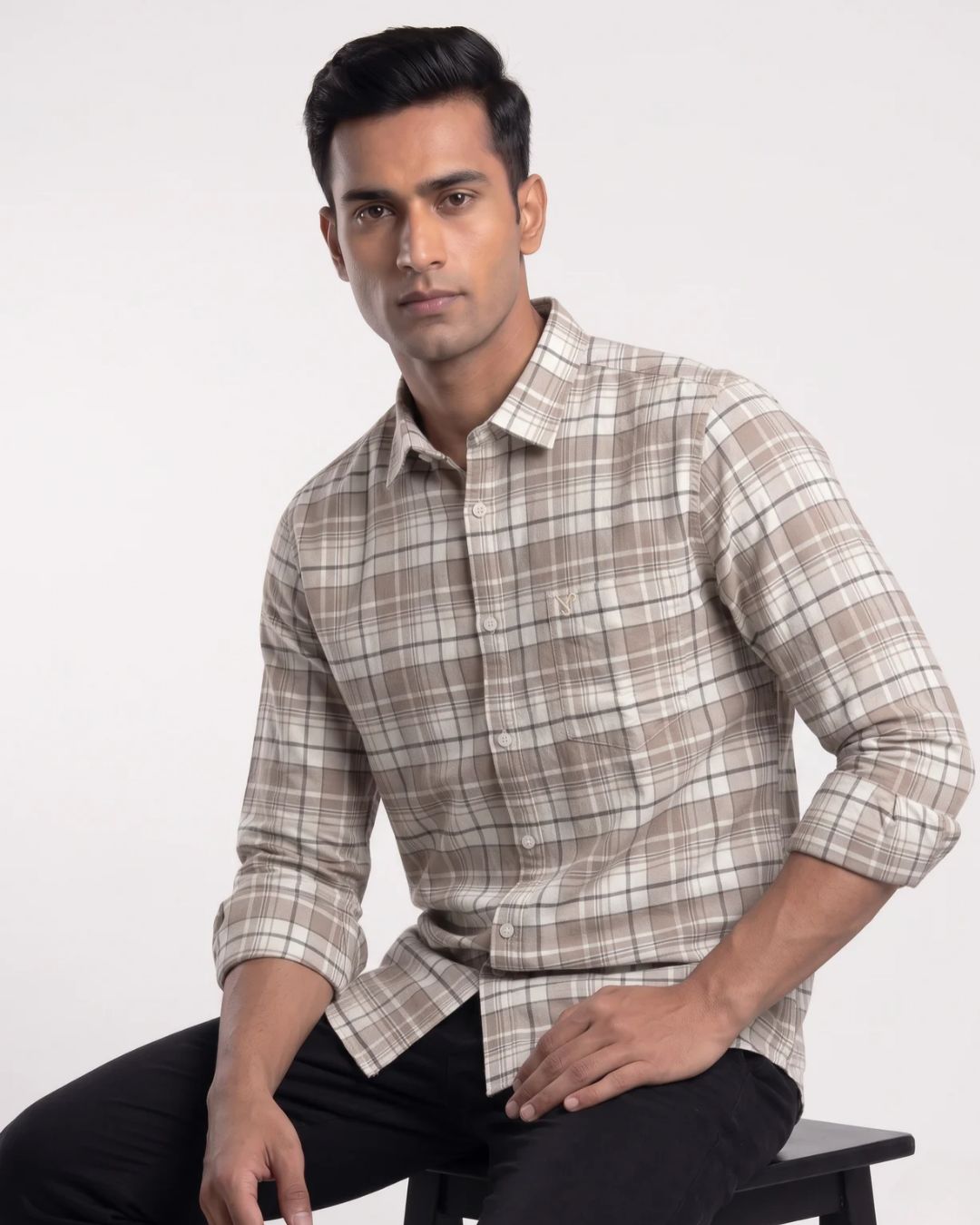 A man wearing a beige and white plaid long-sleeve button-down shirt, sitting on a black stool against a plain light gray background.