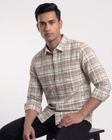 A man wearing a beige and white plaid long-sleeve button-down shirt, sitting on a black stool against a plain light gray background.