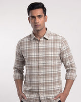 A man wearing a beige and brown plaid long-sleeve button-up shirt, standing against a light gray studio background.