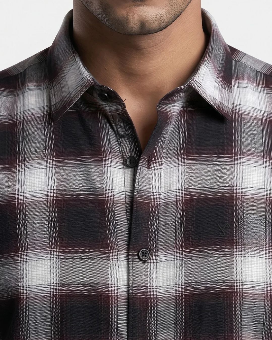 Close-up of a man wearing a plaid button-down shirt in dark gray, white, and burgundy. The shirt features a classic collar, dark buttons, and a subtle embroidered logo on the left chest pocket.