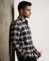 A man in profile wearing a long-sleeve plaid button-up shirt in black, white, and gray, leaning against a textured concrete pillar.
