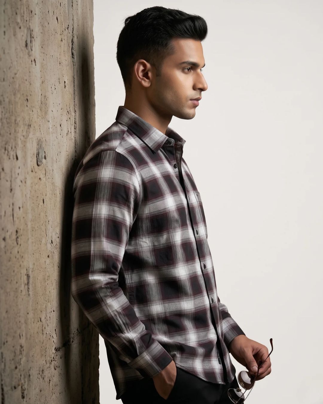 A man in profile wearing a long-sleeve plaid button-up shirt in black, white, and gray, leaning against a textured concrete pillar.
