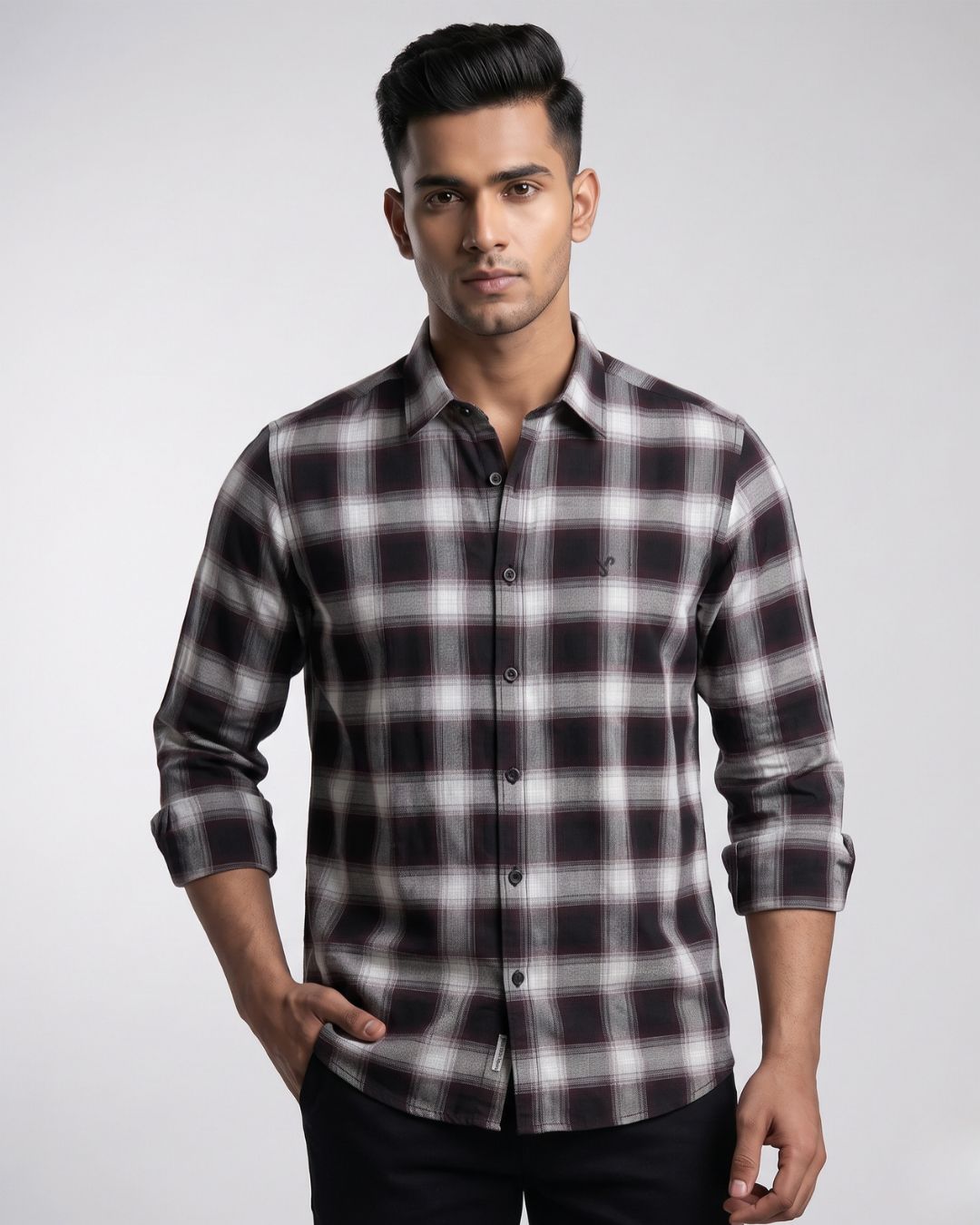 A man wearing a long-sleeve button-up shirt with a black, white, and gray plaid pattern, paired with black trousers, photographed against a light gray studio background.