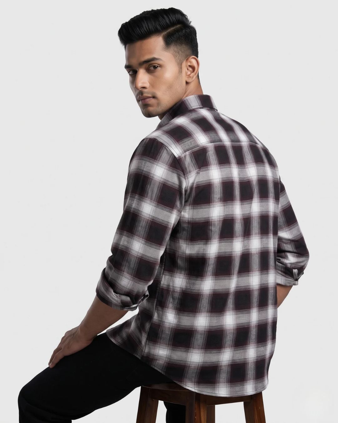 A man wearing a long-sleeve plaid button-up shirt in black, white, and gray, seated on a wooden stool against a light gray background.