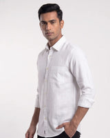 A man wearing a white long-sleeve linen shirt with a chest pocket, shown from the waist up against a plain light gray studio background.
