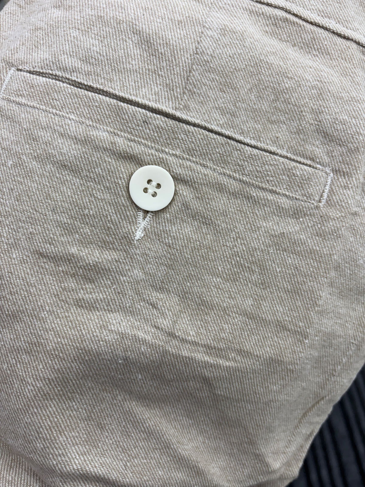 Close-up detail of a light beige textured fabric garment, showing a stitched pocket and a single white button.