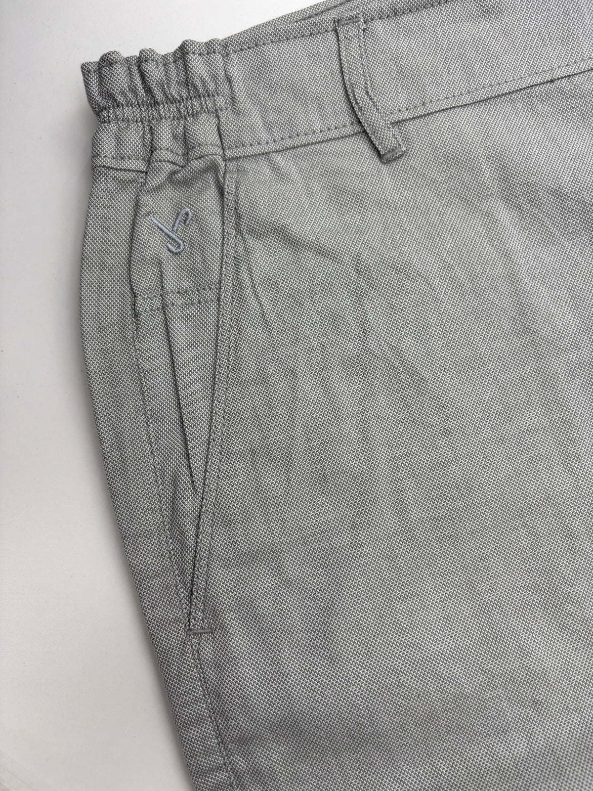 Close-up of light gray textured trousers showing the elastic waistband, belt loops, and side pocket with a small embroidered logo.