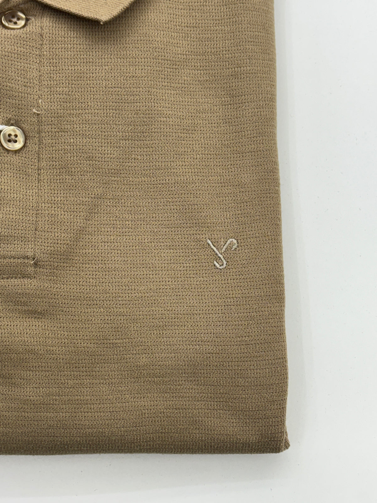 Close-up of a folded tan knit polo shirt showing the collar, button placket, pocket, and a small embroidered logo on a white background.