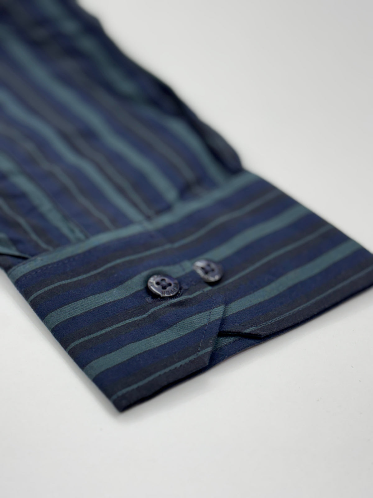 Close-up of a striped dress shirt cuff in navy and teal blue with two dark buttons, photographed on a light gray background.