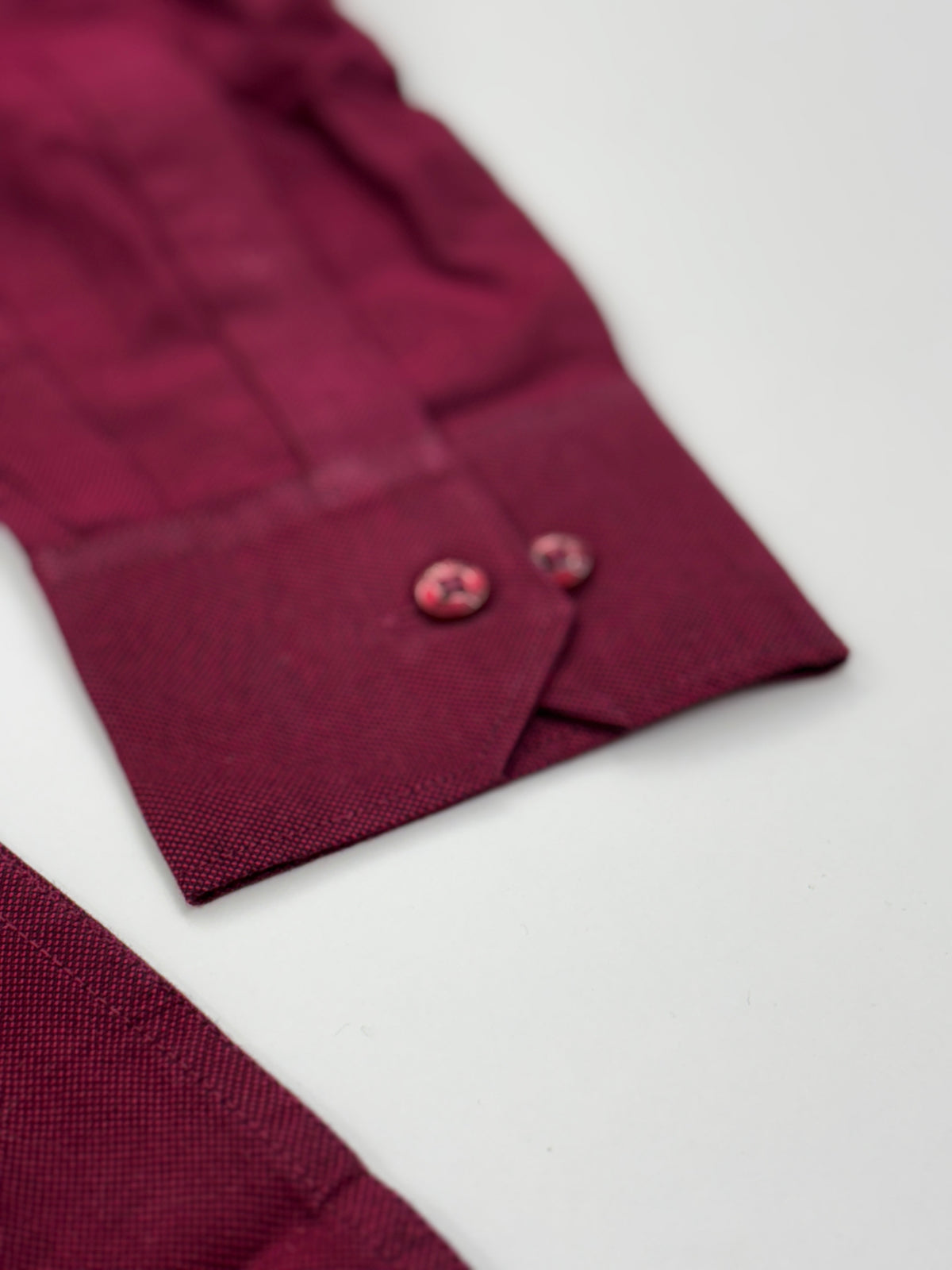 Close-up of a burgundy or deep wine-colored fabric garment, showing the cuff with two matching decorative buttons on a white background.