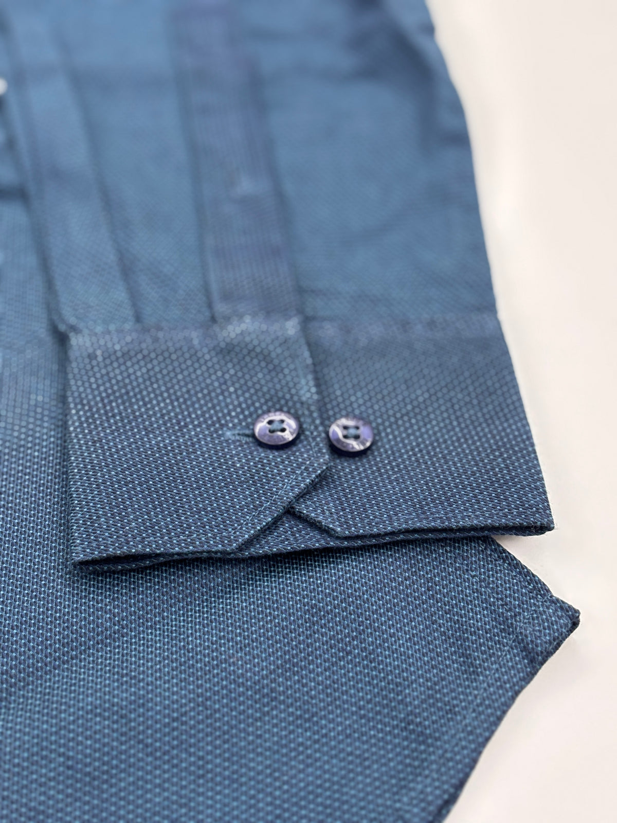 Close-up of a textured blue dress shirt cuff with two buttoned collar tabs, photographed on a light neutral background.