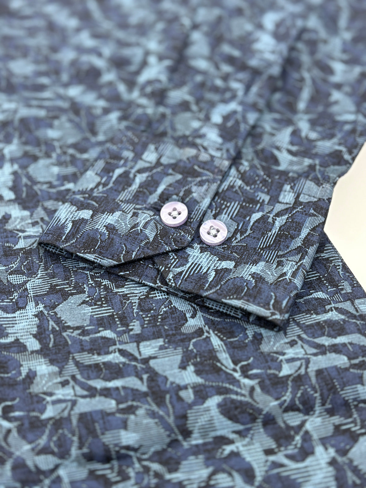 Close-up of a patterned shirt cuff with two light-colored buttons, showing a dark navy and gray abstract floral jacquard fabric.