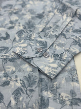 Close-up of a long-sleeve button-up shirt featuring a blue-gray and off-white botanical floral pattern, with a buttoned collar and visible fabric texture.