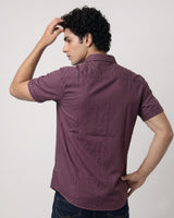 A man wearing a short-sleeve button-up shirt in a dusty plum purple color, shown from the back against a plain light gray studio background.