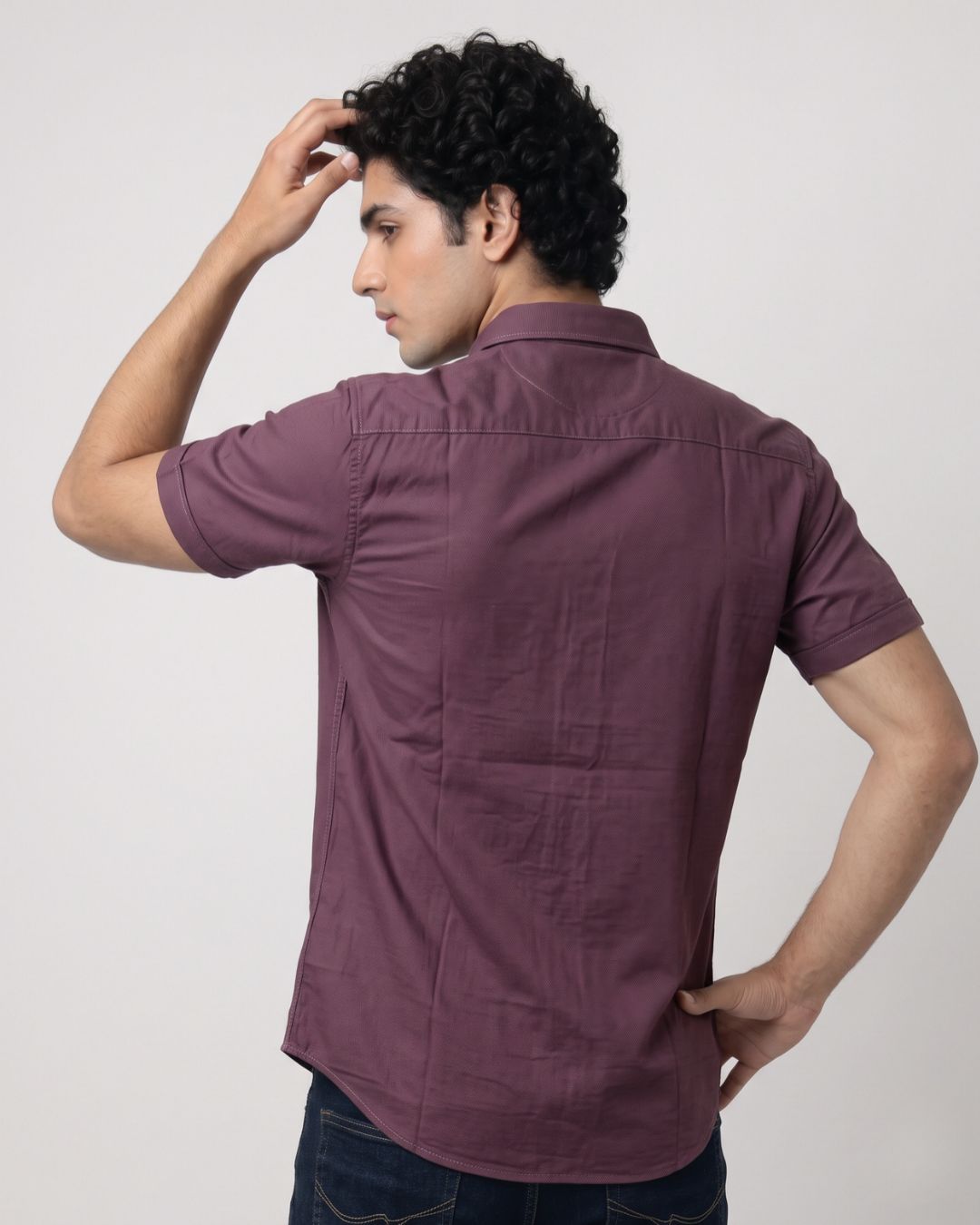 A man wearing a short-sleeve button-up shirt in a dusty plum purple color, shown from the back against a plain light gray studio background.