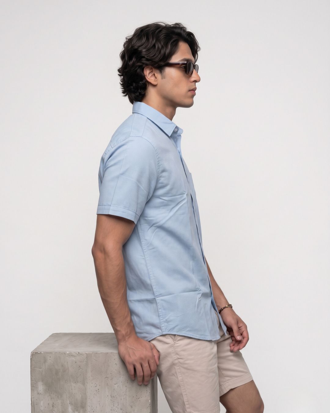 A man wearing a light blue short-sleeve button-up shirt and beige shorts, standing in profile next to a concrete block against a plain white studio background.