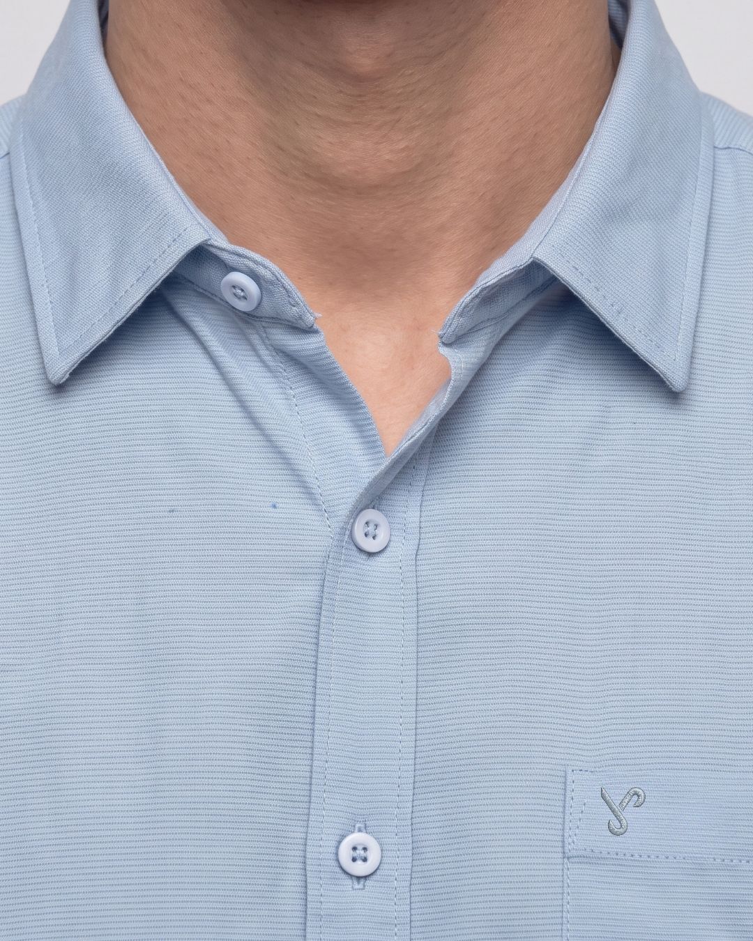 Close-up of a light blue collared button-up shirt worn by a model, showing the collar, button placket, and a small embroidered logo on the chest pocket.