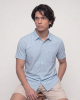 A man wearing a light blue short-sleeve button-up shirt and beige shorts, sitting against a plain off-white background.