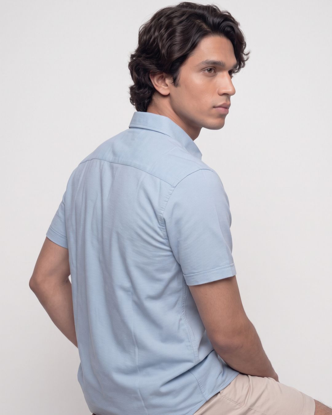 A man wearing a light blue short-sleeve button-up shirt and beige pants, photographed from the back and side against a plain light gray studio background.