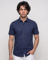 A man wearing a navy blue short-sleeve button-up shirt with a chest pocket, paired with white pants, standing against a light gray studio background.